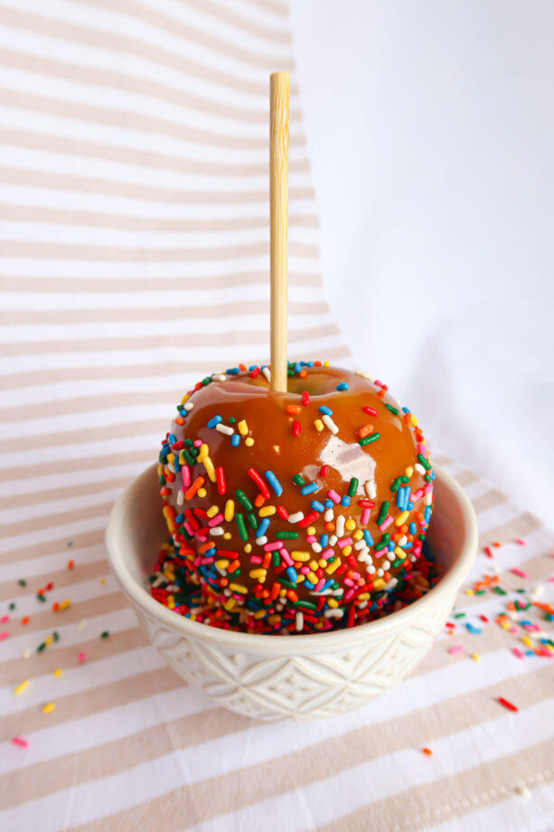 A homemade caramel dipped apple covered in rainbow sprinkles, sitting in a bowl of sprinkles.