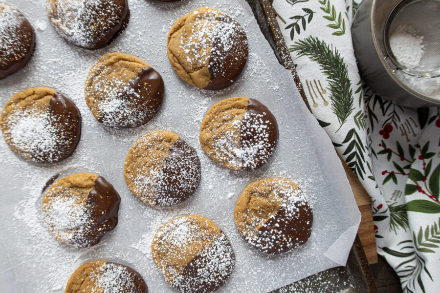 Chocolate dipped gingerbread molasses cookies on a sheet of parchment paper, dusted with powdered sugar. A small bowl of powdered sugar and a holly Christmas napkin sit to the side.