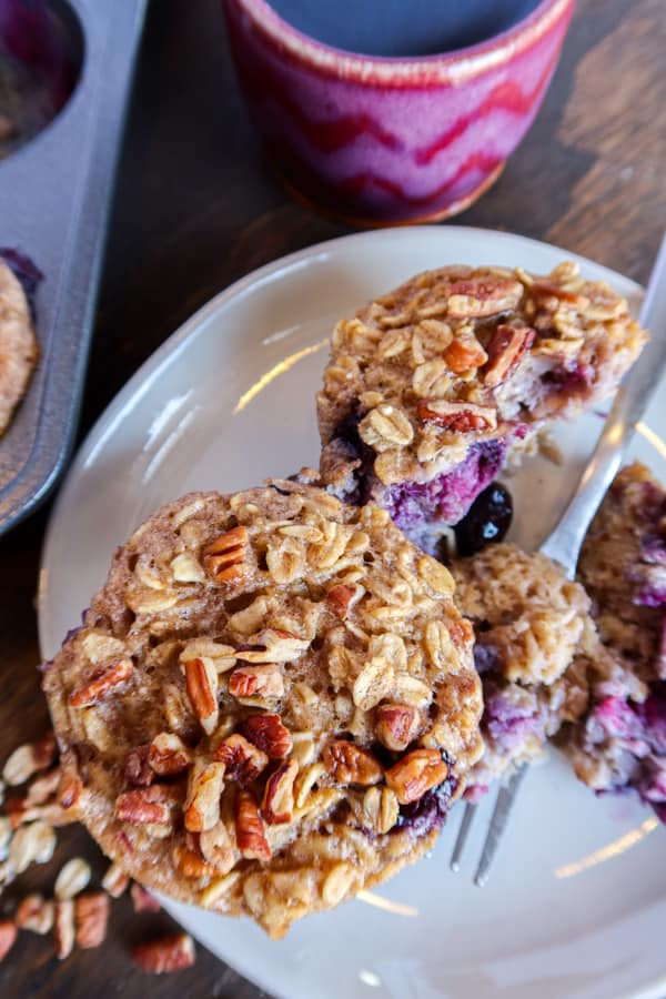 Close up of 2 baked oatmeal cups with blueberries on a white plate with a silver fork. One oatmeal cup is half eaten. A cup of coffee sits to the top of the plate, the muffin pan sits to the left and pecans are scattered to the bottom. 