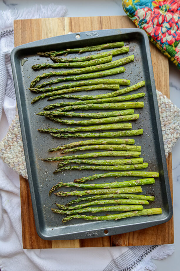Simple, oven roasted asparagus on a dark grey baking sheet, baked and ready to eat! A colorful baking mit sits to the side.