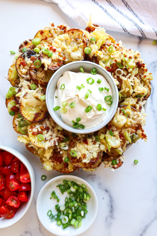 Irish potato nachos stacked around a ramekin full of sour cream on a large, round plate. Nachos and sour cream are sprinkled with chives and a bowl of chives and a bowl of diced tomatoes sit to the side.