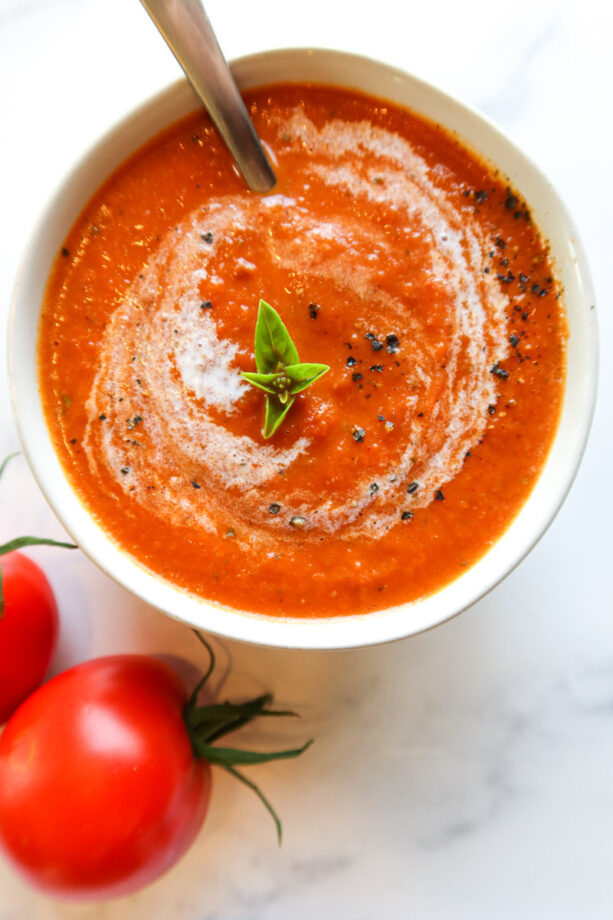 Closeup overhead view shows a small, white bowl of roasted tomato soup with a sprig of basil in the middle. Two small tomatoes sit at the bottom of the bowl. A silver spoon is partially submerged in the bowl of soup.