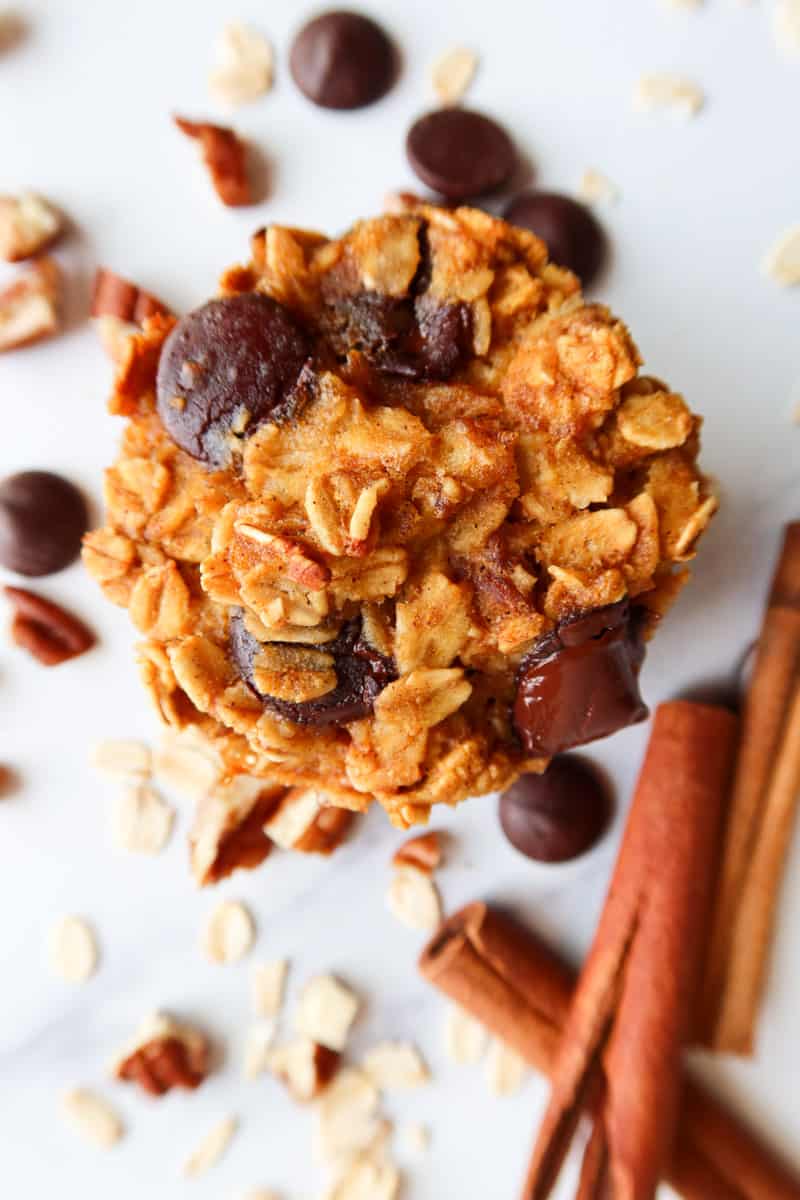 Close up of a pumpkin chocolate chip baked oatmeal cup with cinnamon sticks, chocolate chips, oatmeal and chopped pecans scattered around it.