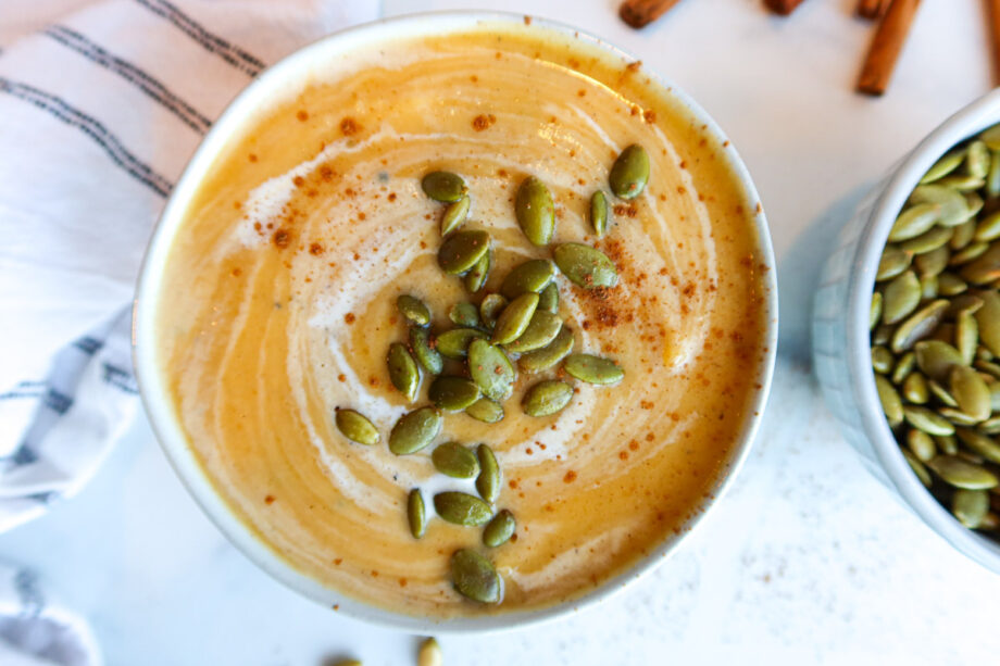 Roasted butternut squash soup in a small white serving bowl. Soup is sprinkled with pumpkin seeds and has cream swirled into it. A little bowl of pumpkin seeds, cinnamon sticks and a black and white striped towel sit to the side.