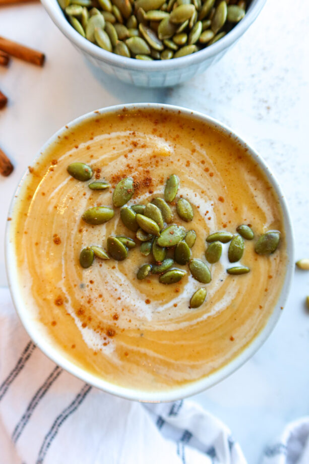 A closeup, overhead view or butternut squash soup in a small white bowl. Soup is topped with roasted pumpkin seeds and a swirl of cream. A small bowl full of pumpkin seeds sits to the top.