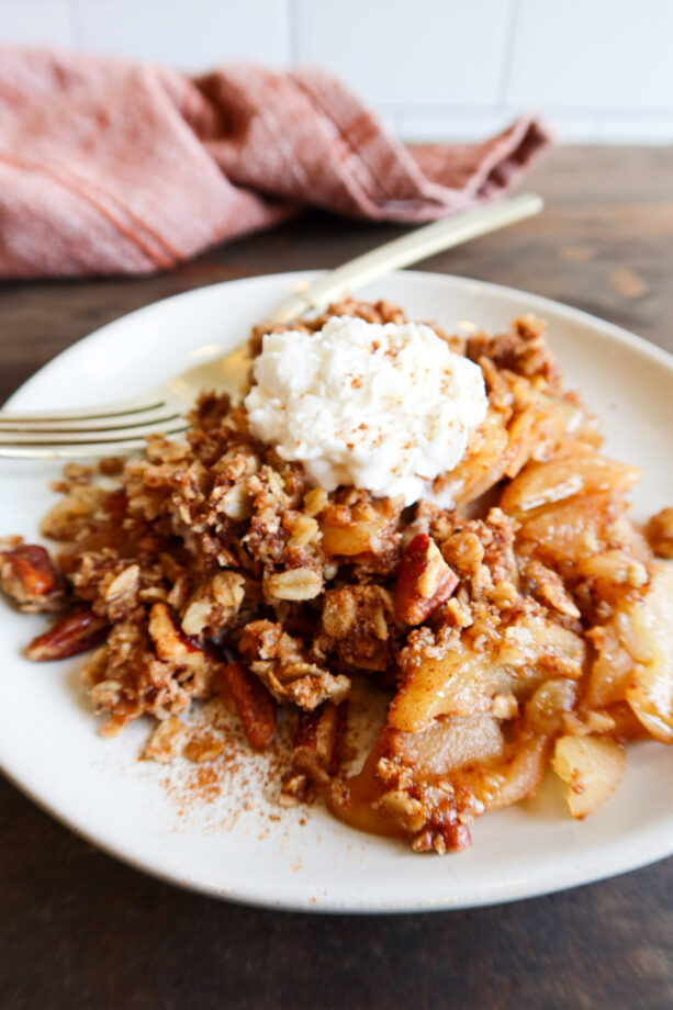 Close up of a small white plate with apple crisp on it. A dollop of whip cream sit on top of the apple crisp and a gold fork sits on the plate.