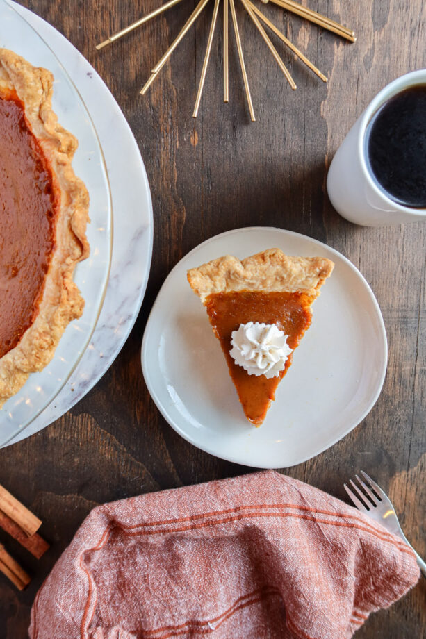 A slice of eggnog pumpkin pie. A red towel, silver fork, coffee, cinnamon stick and the rest of the pie sit to the side.