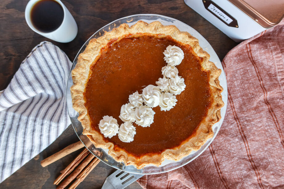 Eggnog pumpkin pie in a glass baking dish. Pie is decorated with whip cream. Cinnamon sticks, a copper and white recipe card, a red towel, a grey striped towel and coffee sit to the side.