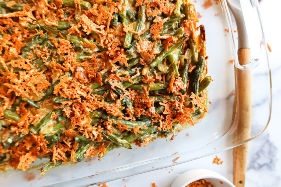 Green bean casserole made from scratch in a large, glass baking dish. Casserole is covered in parmesan cheese crisps. A wooden and grey silicone serving spoon sits to the right and a small bowl of parmesan cheese crisps sits to the bottom. 