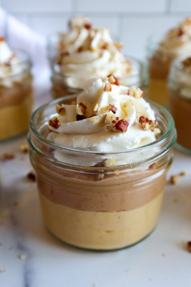 Close up of mini no bake pumpkin chocolate cheesecake jars topped with whipped cream and chopped pecans. Chopped pecan are scattered around the cheesecake jars on the white countertop.