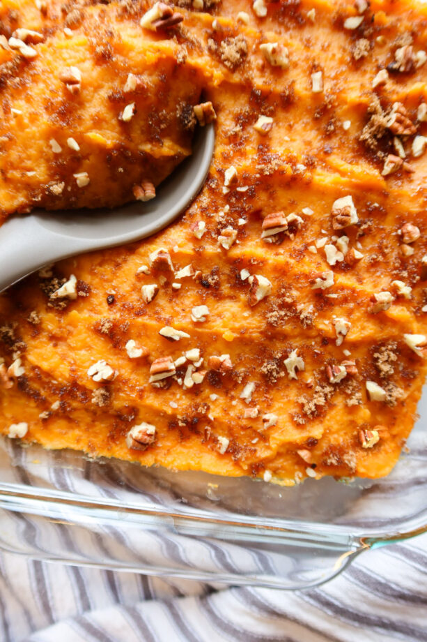 Mashed sweet potatoes (or yams) in a glass baking dish with chopped pecans and a sprinkling of brown sugar on top. A serving spoon has been placed in potatoes and a grey and white striped towel sits below the baking dish.