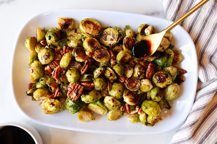 Roasted brussels sprouts mixed with chopped pecans on a white serving plate. A small bowl of balsamic glaze sits to the bottom left. A gold spoon is being used to drizzle glaze on the sprouts. A white and grey striped towel sits to the right. 