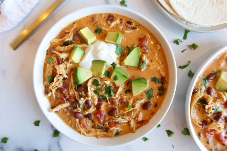 Creamy chicken taco soup in a white bowl with sour cream, chopped cilantro and diced avocado topping.