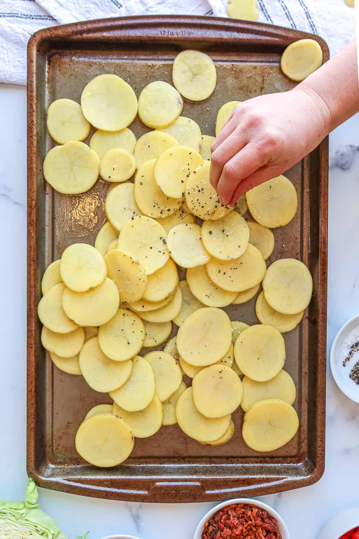 Sprinkling salt and pepper on potato slices atop a baking sheet.