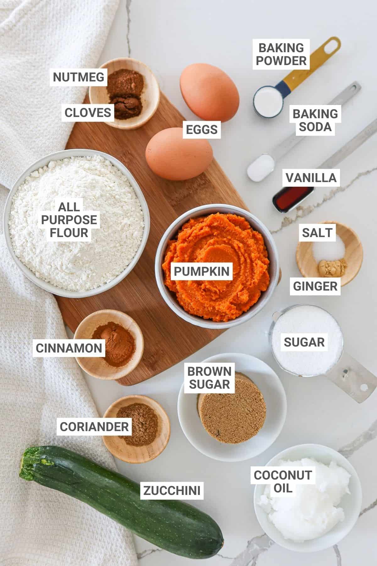 Ingredients for pumpkin zucchini muffins arranged on a white countertop with text overlay labels.