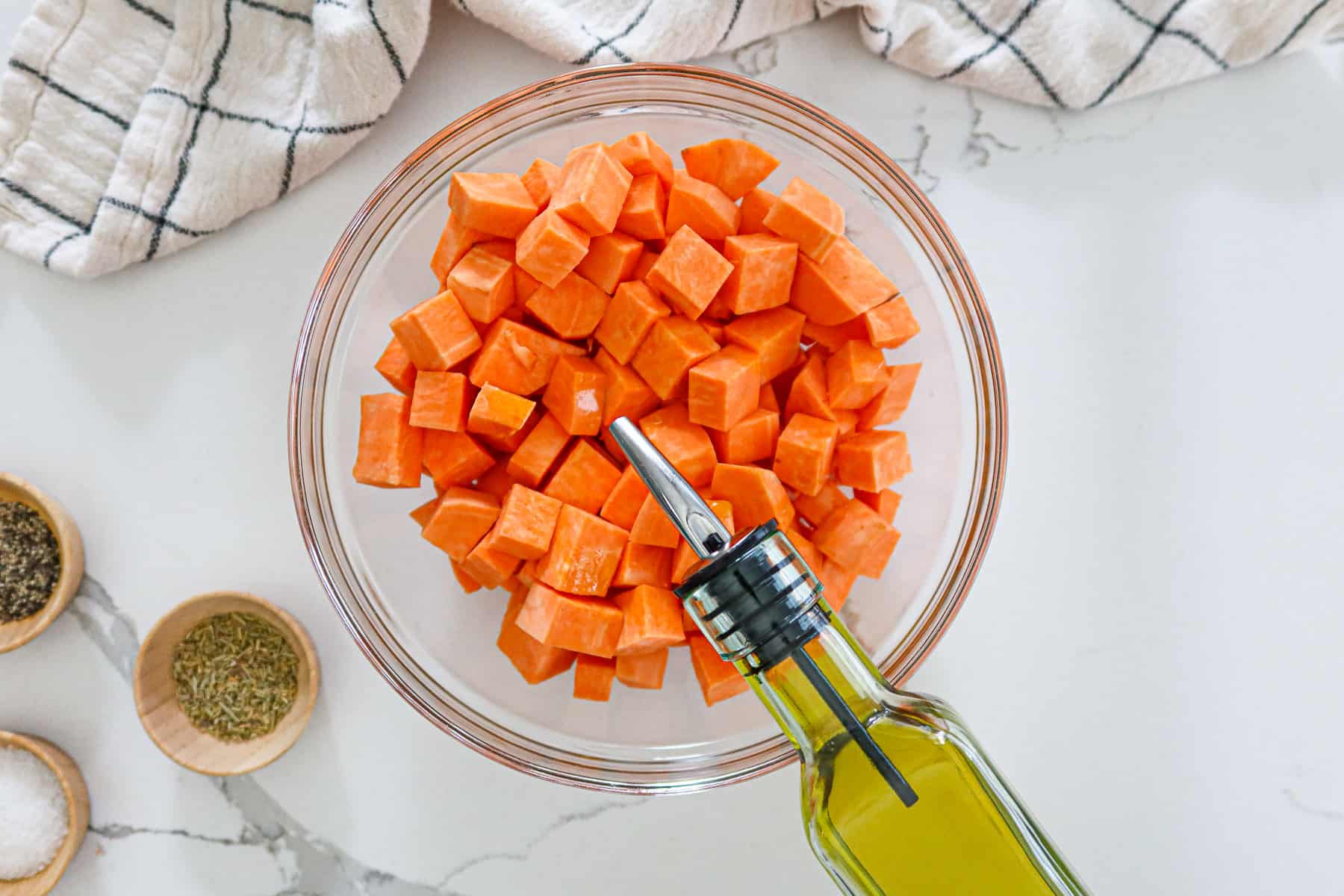 Drizzling olive oil on sweet potato cubes in a glass bowl.