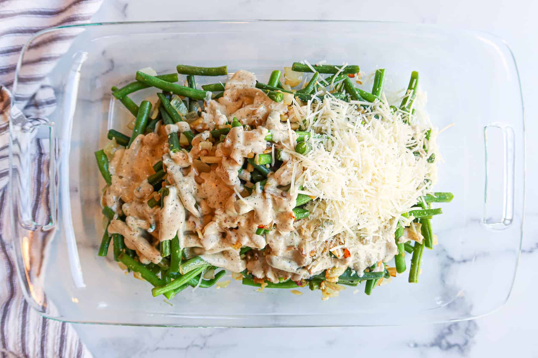 Sautéed green bean mixture in a baking dish covered with sauce and parmesan cheese.