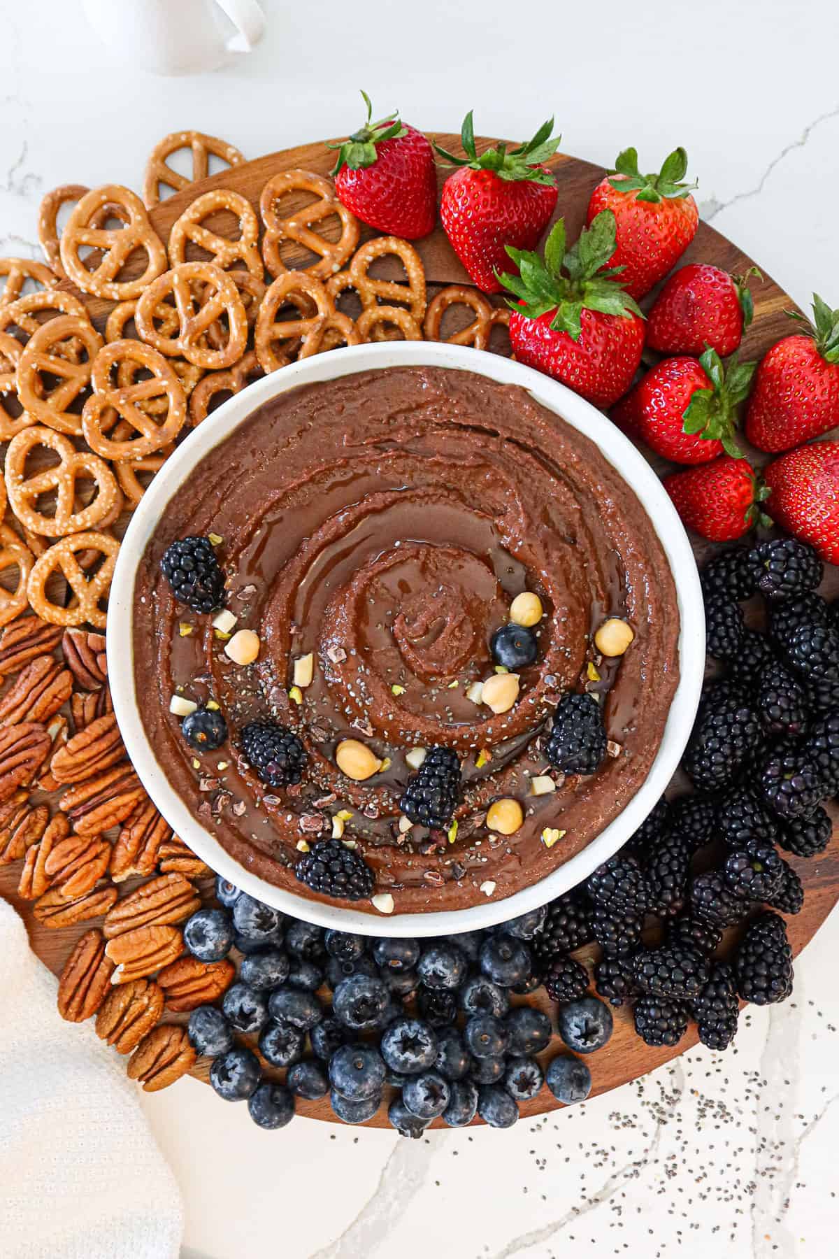 Dark chocolate hummus in a serving bowl surrounded by nuts, pretzels and fresh fruit.