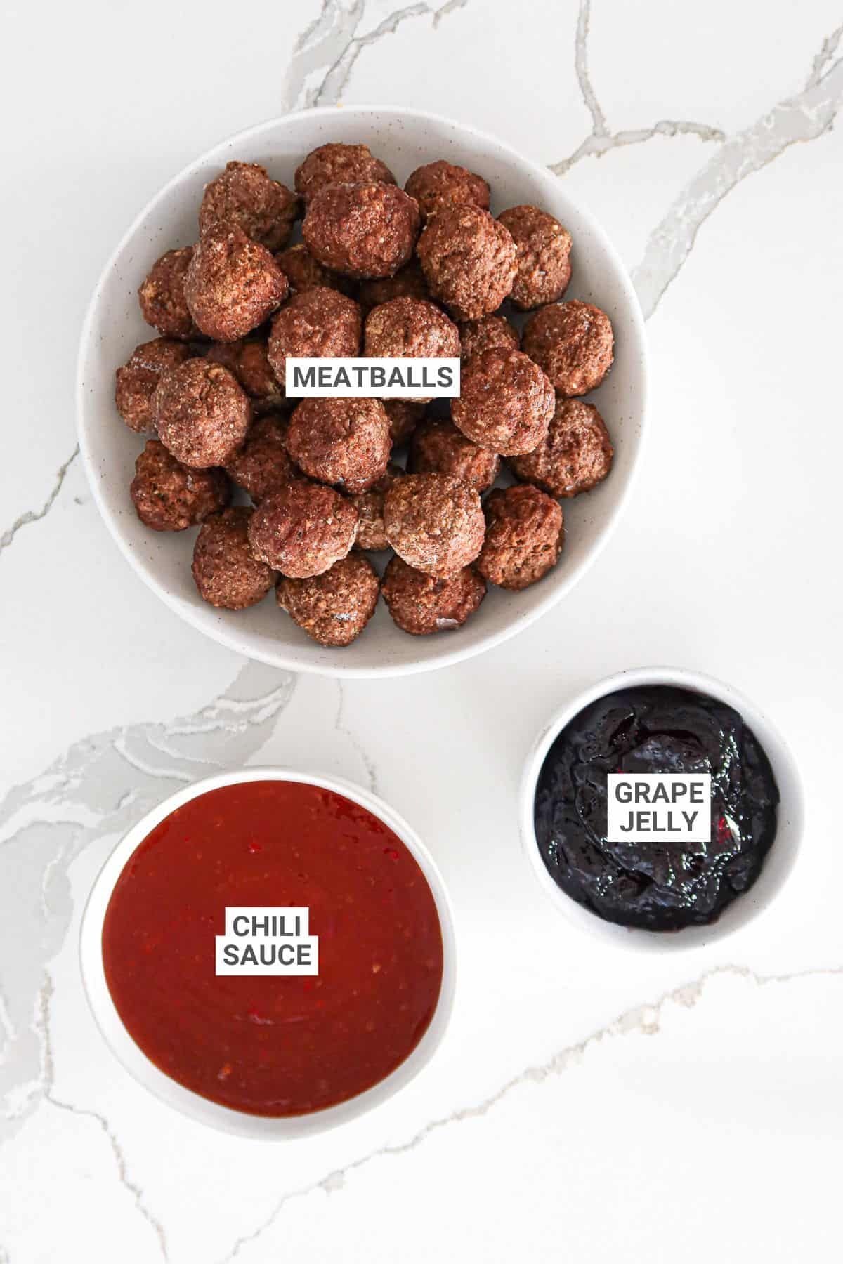 Meatballs, grape jelly, and chili sauce in small bowls on a white countertop.