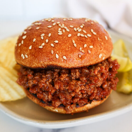 Sloppy joe on a sesame bun with chips and pickles on the side.