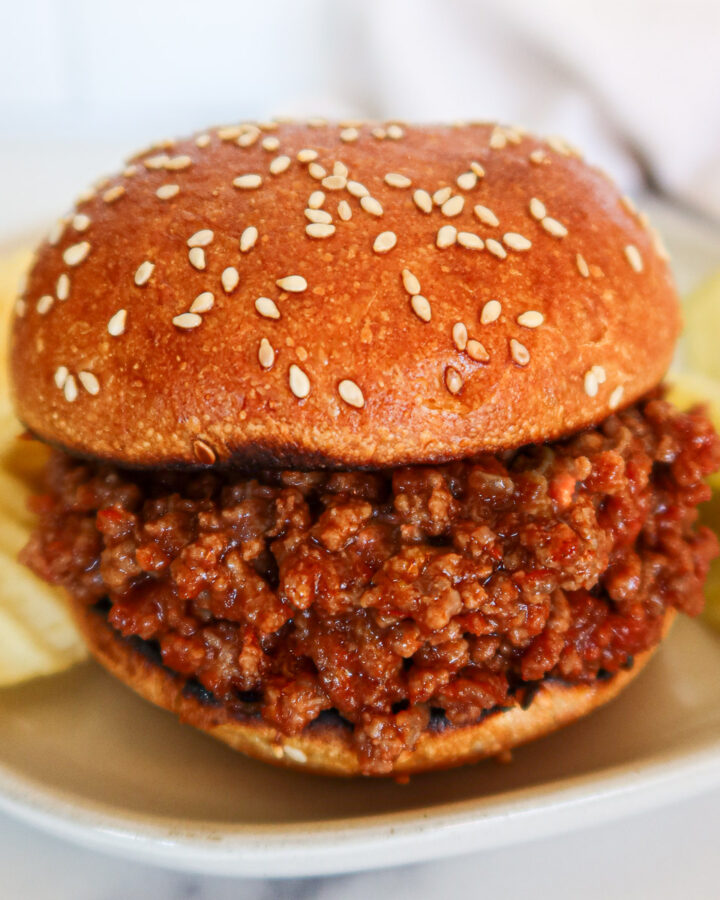 Sloppy joe on a sesame bun with chips and pickles on the side.