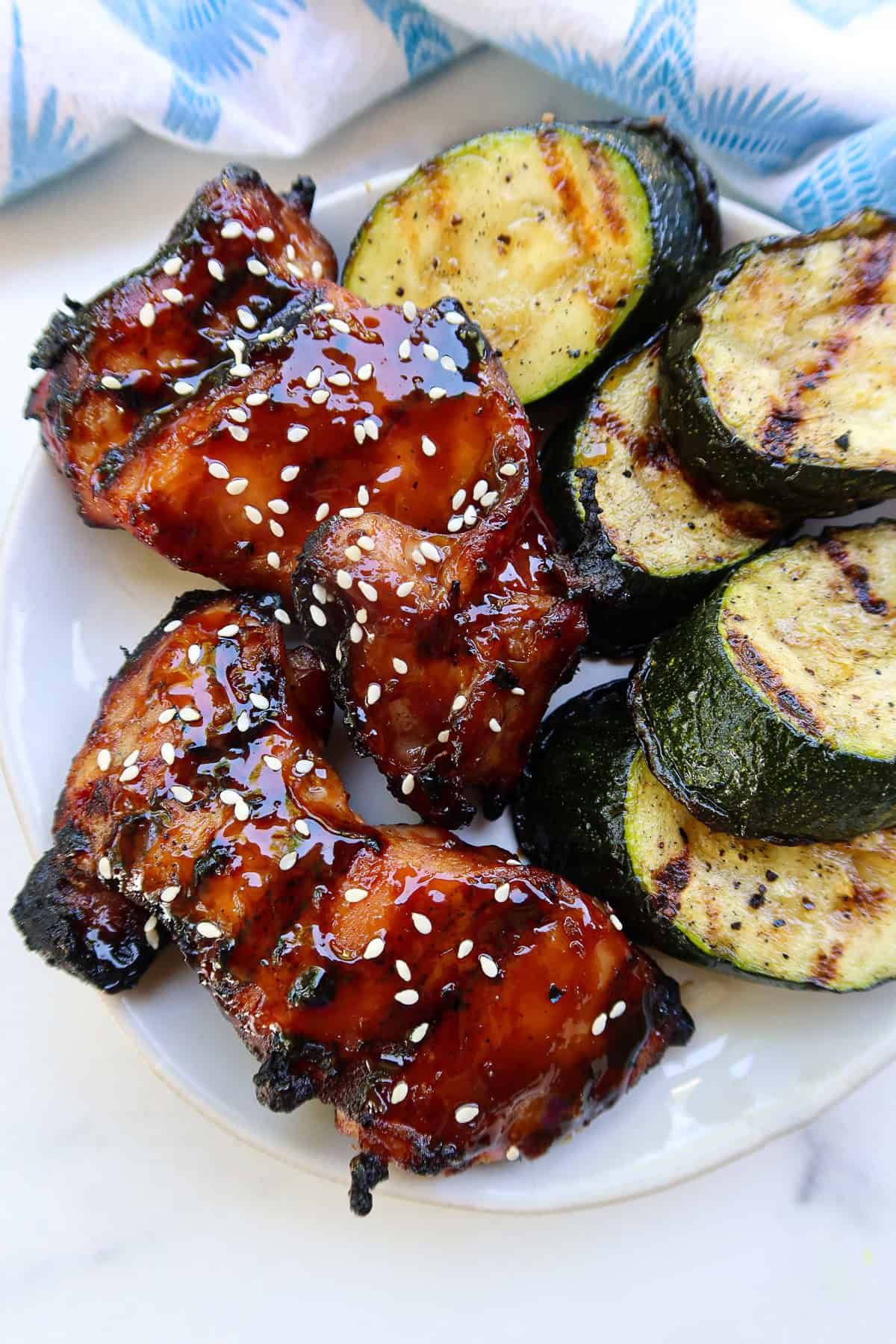 Grilled teriyaki chicken thighs and grilled zucchini on a white plate.